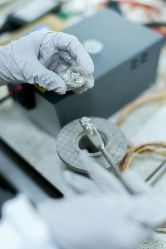 a hand wearing grey gloves handling a grey powder with a tiny spatula