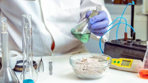 hand wearing purple gloves holding a conical flask filled with green liquid, there are some tools on the bench and a bowl of an off-white chemical in powder form